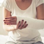 Mid-section of a woman sat down. She has a white medical cast on her left arm which she is supporting with her right hand.